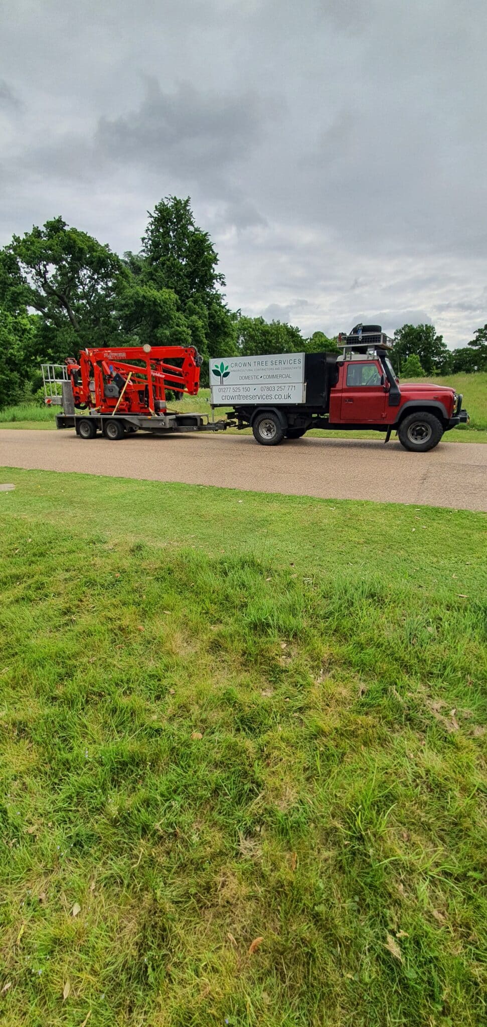 20220525_094505 A red and black lorry with a flatbed trailer carries a piece of red machinery. The lorry has a sign reading "John Tree Services," and is parked on a road with green grass and trees in the background under a cloudy sky.