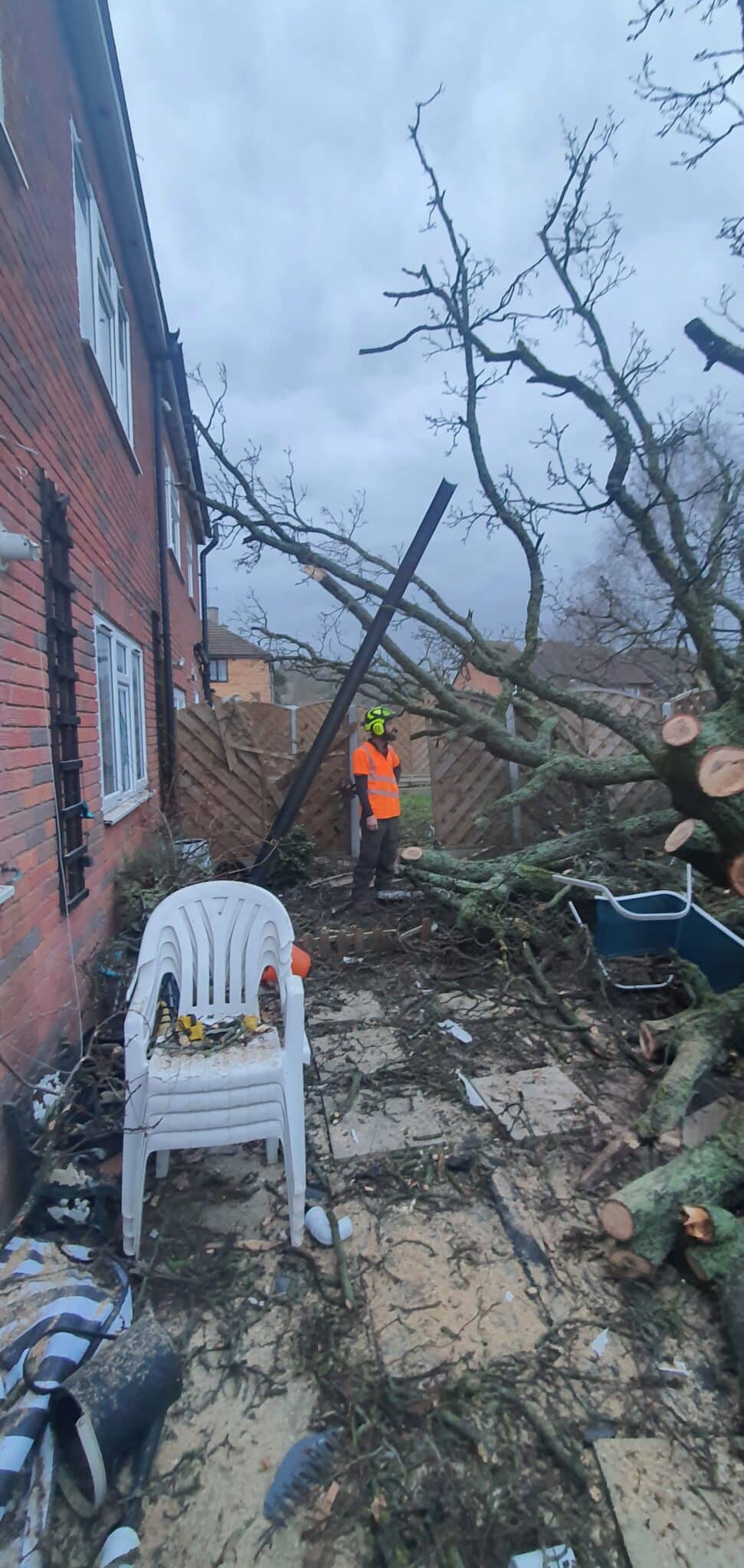 20240207_165252 A worker in an orange vest and helmet stands near a fallen tree that has damaged a back garden, with debris, a broken white chair, and branches scattered near a red-brick house and wooden fence.