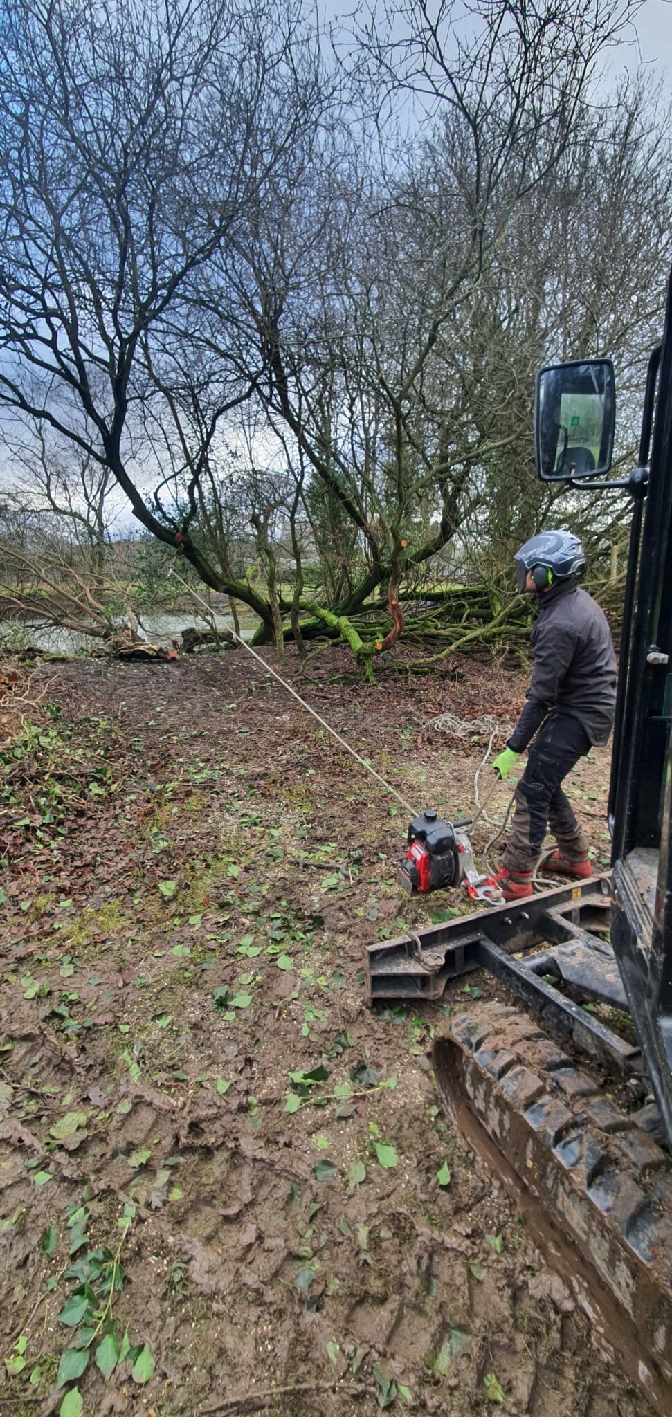 20250107_110743 A person in safety gear operates a winch attached to a fallen tree in a muddy, wooded area, standing next to heavy machinery under a cloudy sky.