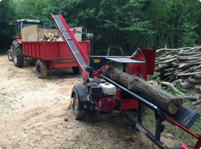 Firefly tree surgeon clearing a large area 395780 A log splitter machine processes a large log, with cut firewood piling up nearby. A red tractor and trailer filled with split wood are parked on a dirt area surrounded by trees.