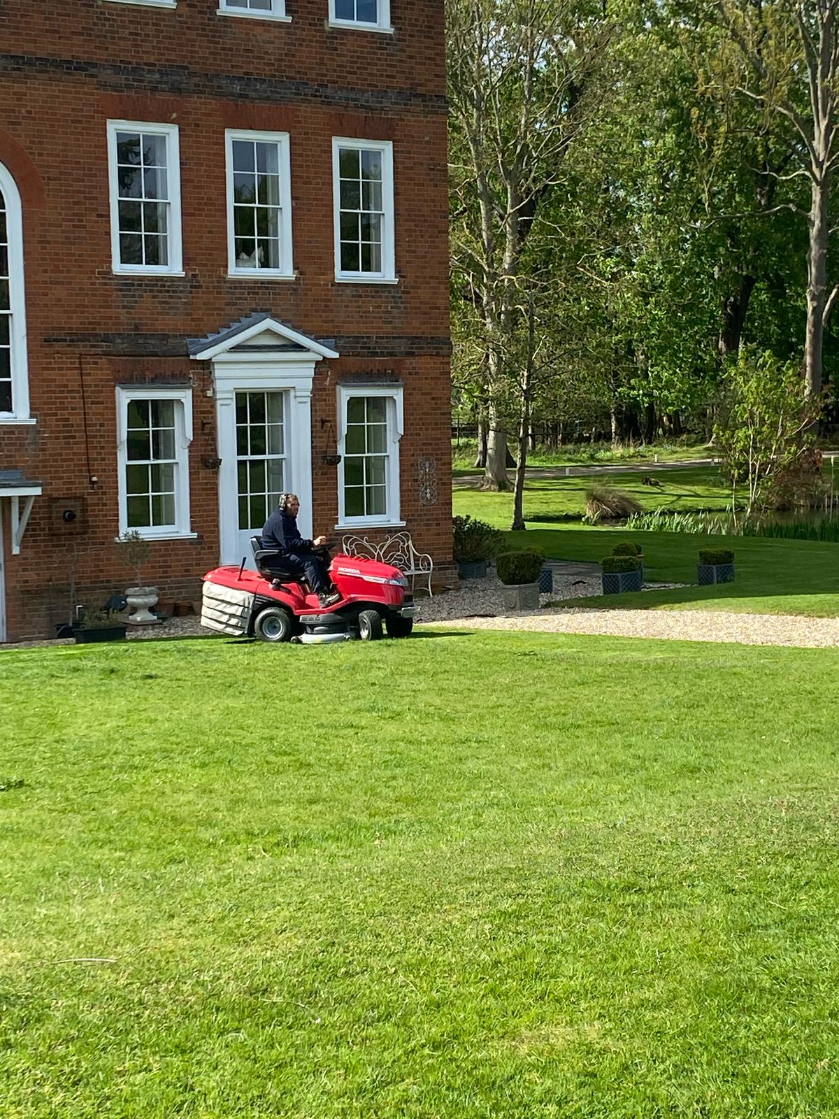 landscape-maintenance-crown-tree-services-essex-2 A person rides a red lawnmower on a green lawn in front of a large brick house with white-framed windows on a sunny day. Trees and garden benches are visible in the background.