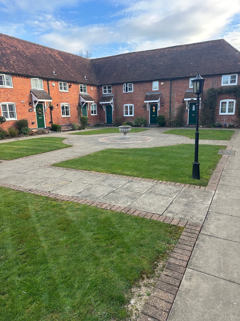 landscape-maintenance-crown-tree-services-essex-3 A courtyard with a circular bench, surrounded by green grass and brick pathways, in front of red-brick terraced houses with green doors and dormer windows. A black lamppost stands near the path.