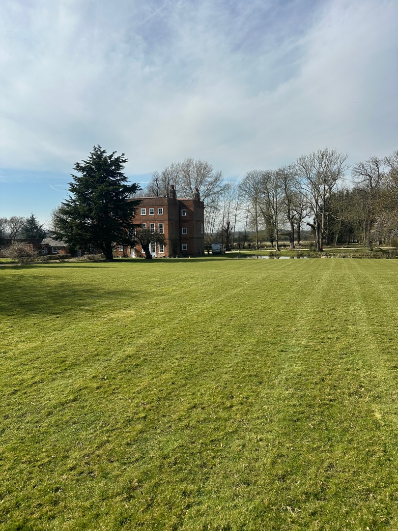landscape-maintenance-crown-tree-services-essex A large maintained green lawn leads to a red-brick house with white windows, partly shaded by a tall evergreen tree. Leafless trees and a blue sky with clouds are visible in the background.