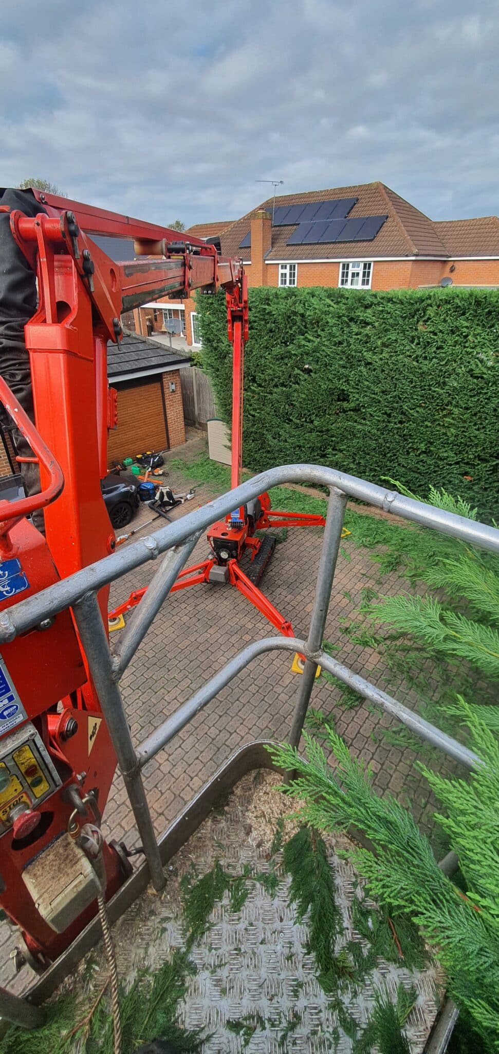 residential-commercial-hedge-trimming-removal-crown-tree View from a cherry picker looking down at a drive with a red lift, hedge, houses, and solar panels on the roof. Tree branches and metal railings are visible in the foreground.