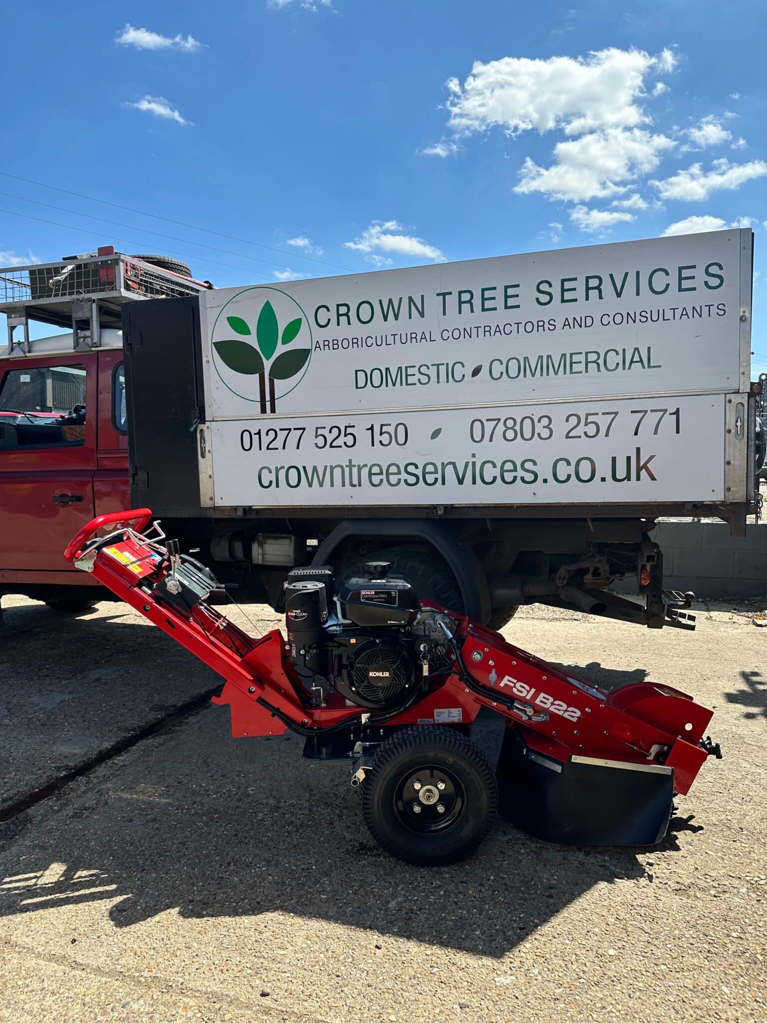 tree-stump-removal-grinding-crown-tree-services A red stump grinder is parked in front of a Crown Tree Services lorry, which displays contact information and a green leaf logo on its side, under a sunny sky.