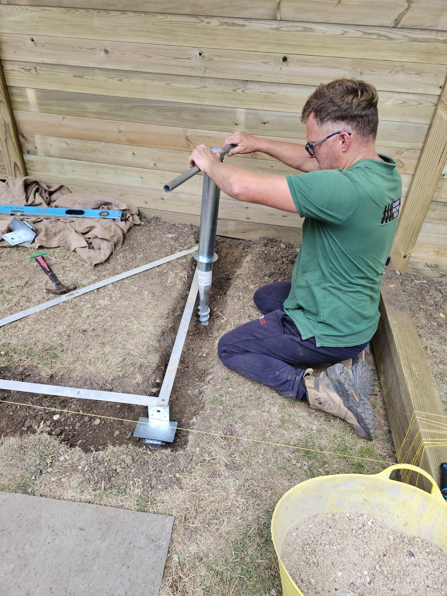 arboricultural-supervision-services A man kneels on the grass using a tool to install a metal ground screw next to a wooden fence, with construction materials and tools scattered nearby.