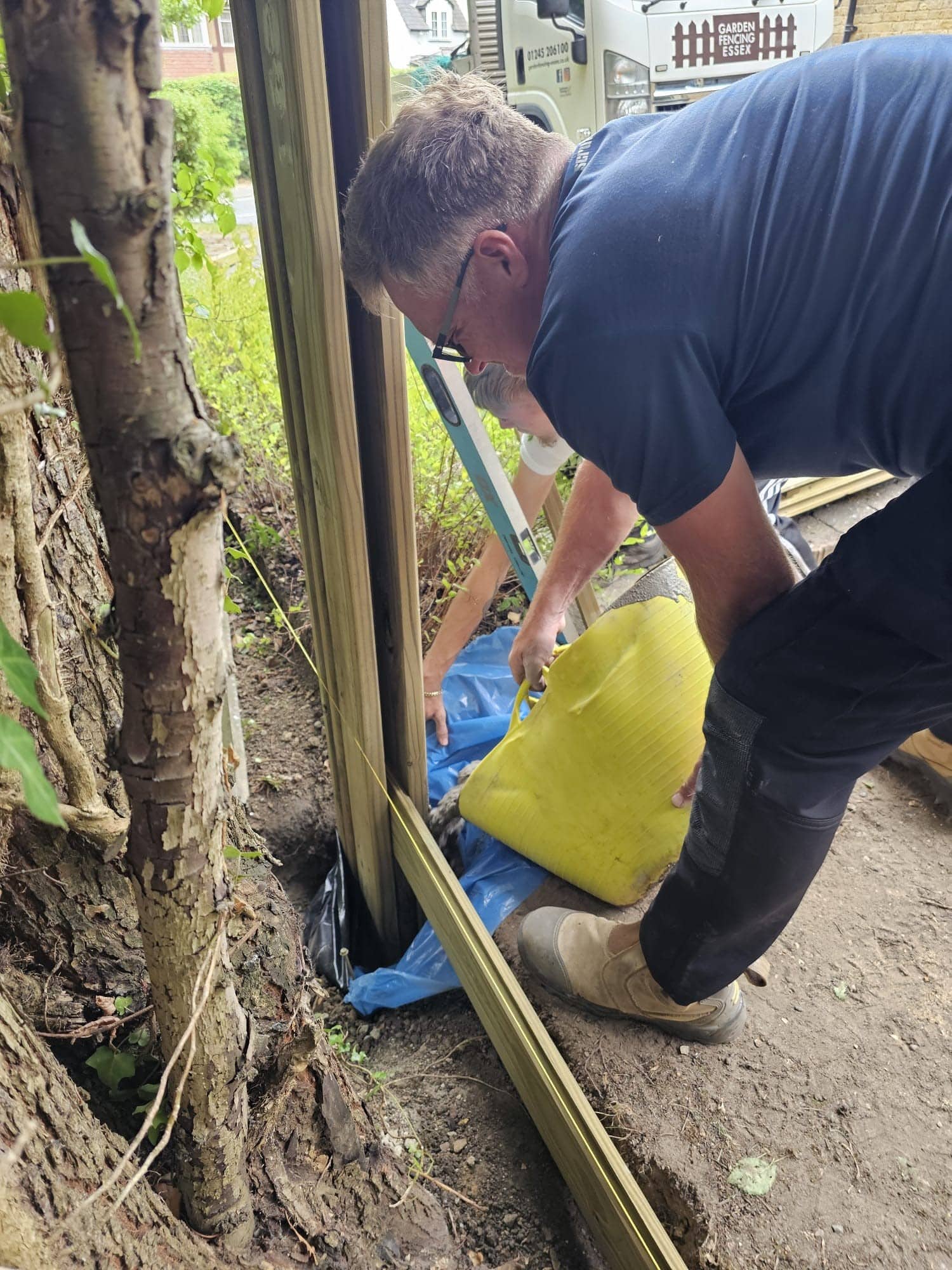 crown-tree-arboricultural-supervision-services A man wearing glasses pours a yellow bag of material, possibly cement, into a hole next to wooden fence posts whilst working outdoors beside trees and a garden.