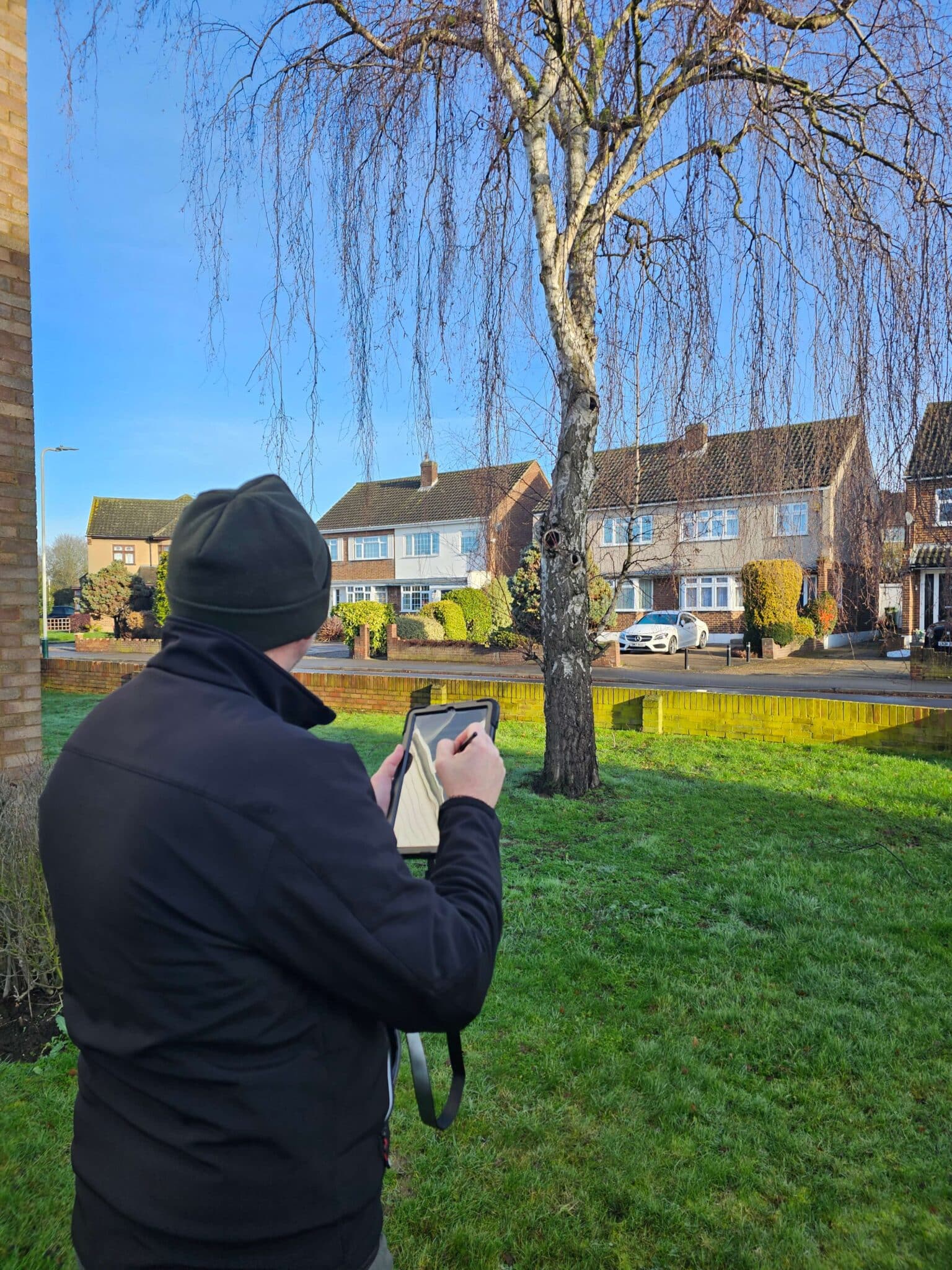 A person in a black jacket and beanie stands on a grassy lawn, using a tablet or clipboard to take notes or draw, with suburban houses and a bare tree visible in the background.
