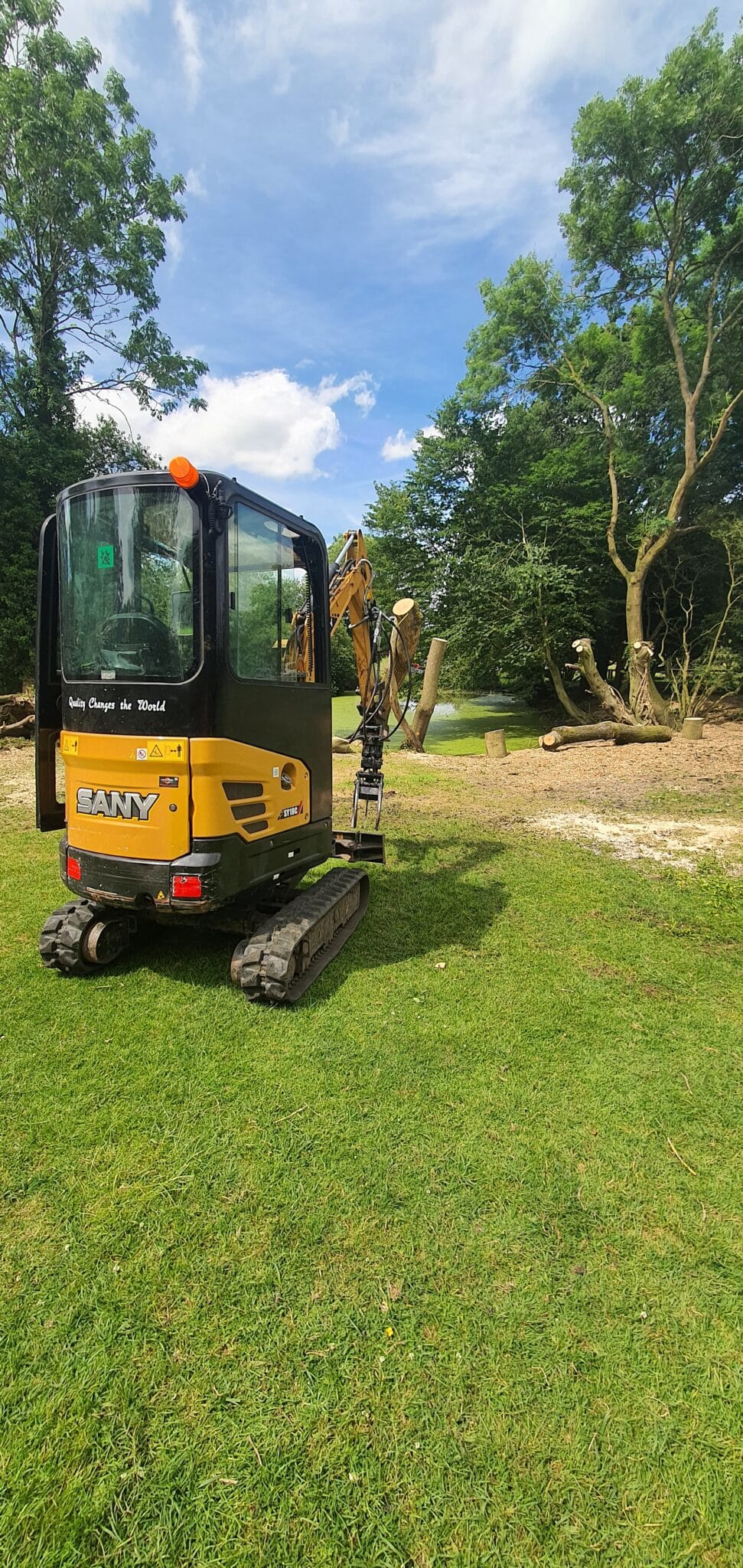 A small yellow SANY excavator is parked on green grass near trees under a partly cloudy sky, with chopped logs and branches visible in the background.