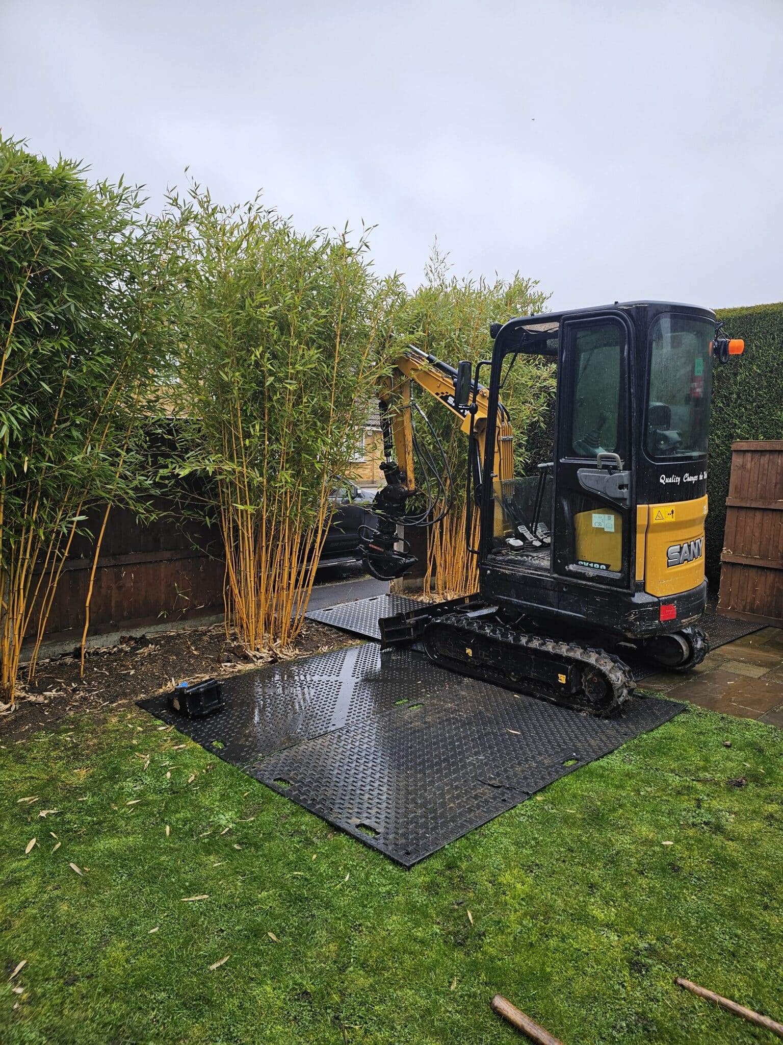 A small black and yellow digger is removing tall bamboo plants from a garden, with protective mats placed on the grass to prevent damage. The scene is overcast and the garden is bordered by wooden fencing and hedges.