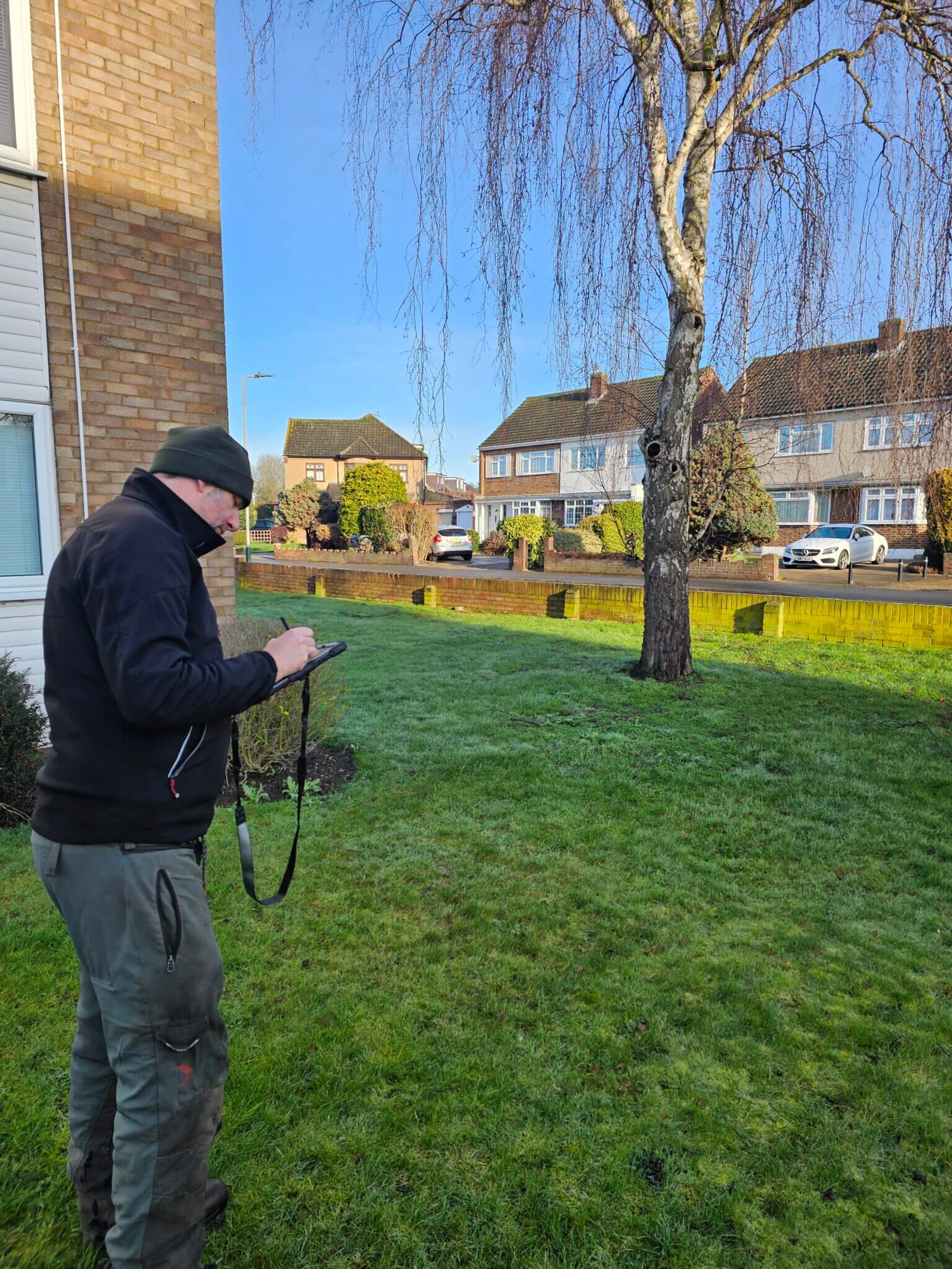 A person wearing a hat and dark jacket stands on a grassy lawn using a tablet, next to a tree and a residential building. Houses and cars are visible across the road under a blue sky.